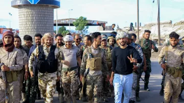 La imagen muestra a un grupo de hombres, aparentemente soldados, caminando juntos por una calle. Algunos llevan uniformes militares y portan armas, mientras que otros visten ropa más casual. Hay un ambiente de camaradería, y uno de los hombres en el centro hace un gesto de paz con la mano. El fondo muestra un paisaje urbano y estructuras que parecen ser parte de una ciudad en reconstrucción o conflicto. La escena refleja un momento de unidad y posiblemente celebración.