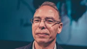 Homme souriant avec des lunettes, cheveux courts et gris, fond flou en arrière-plan.