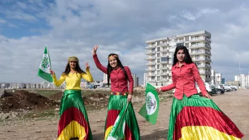 L'image montre trois femmes portant des jupes colorées, affichant des drapeaux et symboles. Elles sont entourées d'un paysage urbain, avec des bâtiments en arrière-plan et un terrain dégagé. Les femmes semblent célébrer un événement, affichant des sourires et des gestes de victoire. Les couleurs vives de leurs vêtements et des drapeaux évoquent un sentiment de fierté culturelle.