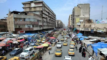 L'image présente une scène urbaine animée, probablement dans une grande ville. On peut voir une rue bordée de bâtiments qui semblent vieux et usés. La circulation est dense, avec de nombreux taxis jaunes qui circulent dans les deux sens. Des échoppes et des marchés de rue sont visibles, avec des auvents colorés, indiquant une ambiance de commerce local. Les gens marchent le long des trottoirs, et l'ensemble de la scène donne une impression de mouvement et de vie citadine. Le ciel est partiellement nuageux, ce qui suggère un climat chaud.