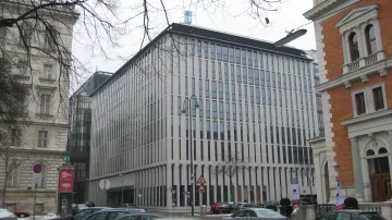 L'image montre un bâtiment moderne avec une façade composée de lignes verticales blanches. Il se trouve à proximité d'autres structures architecturales, dont l'une semble plus ancienne, en pierre avec des ornements. Le paysage est urbain et il y a plusieurs voitures garées au premier plan. Le sol est couvert de neige, ce qui suggère une ambiance hivernale. Le ciel est nuageux, ajoutant à la scène une atmosphère sobre.