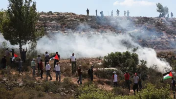 L'image montre une scène de manifestation en plein air. Au premier plan, plusieurs personnes portent des drapeaux palestiniens, tandis que d'autres semblent être en train de marcher ou de s'arrêter. Il y a une atmosphère de tension, avec de la fumée s'élevant dans l'air, probablement due à des gaz lacrymogènes ou à un autre type de dispersion de foule. En arrière-plan, on aperçoit des personnes positionnées sur une colline, qui semblent observer la situation. Le paysage est naturel, avec des arbres et des terrains rocheux.