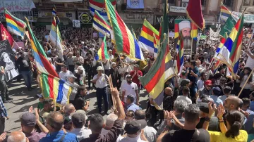 L'image montre une grande foule rassemblée lors d'une manifestation. Les participants tiennent des drapeaux colorés, probablement de différentes communautés ou groupes. On peut voir des visages de personnes connues affichés sur des pancartes. L'atmosphère semble dynamique et engagée, avec des manifestants applaudissant et exprimant leurs opinions. Le décor urbain suggère que l'événement se déroule dans une ville animée.