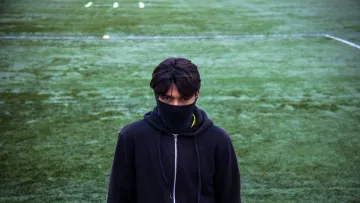 The image features a young person standing on a green sports field, possibly a soccer pitch. They are wearing a black hoodie and have a black mask covering the lower part of their face. The focus is on their intense gaze, and they appear to be looking directly at the camera. The field is marked with white lines and has some yellow cones in the background, suggesting a training or practice session. The overall mood is somewhat mysterious and serious.