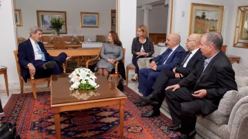 L'image montre un groupe de personnes assises dans un salon bien éclairé et décoré. Il y a une table basse au centre avec un arrangement floral blanc. À gauche, une personne est assise dans un fauteuil, écoutant attentivement. Les autres personnes sont disposées autour de la table, portant des costumes formels et engageant une conversation. Les murs sont ornés de tableaux, et le décor évoque une atmosphère professionnelle et conviviale.