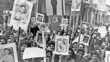 L'image montre une foule de personnes rassemblées dans une rue, tenant des pancartes et des portraits en protestation. On peut voir un dessin d'une potence au centre de l'affichage, suggérant un contexte de tension ou de révolte. Les visages des manifestants expriment diverses émotions, et l'environnement semble animé. Des palmiers sont visibles en arrière-plan, ajoutant une touche de verdure à la scène.