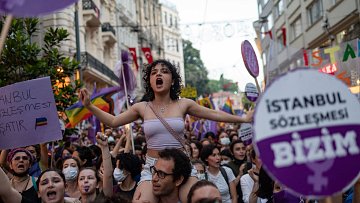 L'image montre une foule lors d'une manifestation, avec des personnes brandissant des pancartes et des drapeaux, notamment des drapeaux arc-en-ciel. Au centre, une jeune femme exprime sa détermination en chantant ou en criant, tandis qu'elle est portée par une autre personne. Tout autour, les manifestants portent des vêtements aux couleurs violettes et blanches, et l'atmosphère semble énergique et engagée. Les pancartes mentionnent des références à des droits, notamment le "contrat d'Istanbul", qui met en avant des thèmes de protection et d'égalité.