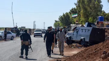 تصویر، یک خیابان با خودروها و کامیون‌ها است. چند نفر با لباس نظامی در حال حرکت هستند. دیوارها و درختان اطراف هستند.