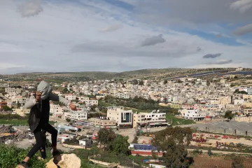 تظهر الصورة مشهداً طبيعياً لمدينة أو قرية، حيث تطل المباني السكنية على مساحات خضراء. في مقدمة الصورة، يوجد شخص يقف على صخرة، يبدو أنه في حالة حركية، بينما تظهر خلفه منظر عام للمدينة والمناطق المحيطة بها. السماء مليئة بالغيوم، مما يعكس أجواء متغيرة. تبرز في الصورة شوارع ومزارع، مما يعطي انطباعاً عن البيئة المحلية.