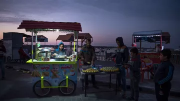 L'image montre un stand de nourriture de rue à la tombée de la nuit, avec un ciel coloré par le crépuscule. Au premier plan, un vendeur prépare des aliments, tandis que plusieurs jeunes clients attendent. Le stand est décoré avec des affiches et contient des produits alimentaires, tels que des pâtisseries. En arrière-plan, on peut apercevoir d'autres stands illuminés et des chaises, créant une ambiance conviviale.