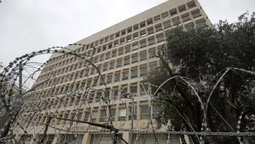 الصورة تظهر مبنى عالٍ يظهر بشكل معماري حديث، ويتواجد في المقدمة سياج من الأسلاك الشائكة. السماء فوق المبنى ملبدة بالغيوم، مما يوحي بجو قاتم. تحيط بالمبنى بعض الأشجار، مما يضيف لمسة خضراء في المشهد. تعكس الصورة شعورًا بالأمان أو القيود، نظراً للسياج الذي يحيط بالمكان.