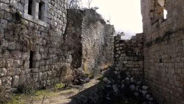 L'immagine mostra un paesaggio di rovine in un'area collinare. Ci sono muri di pietra, molti dei quali sembrano in stato di abbandono e degrado. La vegetazione è presente, con erbacce e piante che crescono tra le pietre. La luce che filtra suggerisce un'atmosfera di tranquillità, ma anche di desolazione, evidenziando il contrasto tra la natura e i resti architettonici. Nel complesso, il luogo appare come un residuo di una civiltà passata, avvolto in un silenzio nostalgico.