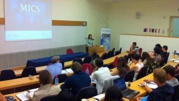 L'image montre une salle de classe ou un auditorium où une présentation est en cours. À l'avant, une intervenante se tient près d'un pupitre, probablement en train de s'adresser à l'auditoire. En arrière-plan, un écran affiche le mot "MICS" accompagné d'une carte du monde. Le public, composé d'une douzaine de personnes, semble attentif et prend des notes sur des ordinateurs portables ou des bloc-notes. La disposition des sièges est en rangées, et quelques personnes sont assises avec des tasses et des boissons. L'ambiance semble académique et interactive.