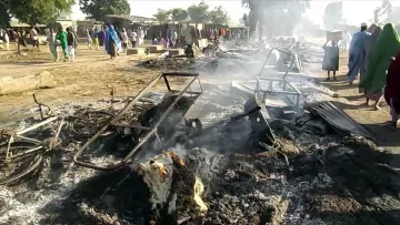 La imagen muestra un escenario devastador después de un incendio. Hay estructuras de metal chamuscadas y escombros humeantes en el suelo. En el fondo, algunas personas parecen estar observando la escena, mientras que el humo se eleva en el aire. El paisaje refleja una atmósfera de destrucción, posiblemente en un área rural o un mercado.