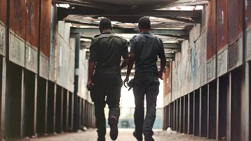 L'image montre deux hommes marchant côte à côte dans un passage couvert. Ils portent des uniformes de couleur sombre et se déplacent vers la sortie, avec leurs silhouettes se détachant contre la lumière. Les murs du passage semblent usés, avec des graffitis visibles, et le sol est pavé. L'atmosphère est à la fois urbaine et légèrement mélancolique, donnant une impression d'intimité entre les deux personnages.