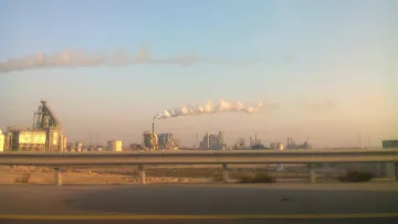 The image depicts an industrial landscape with several factories. In the foreground, there is a highway barrier, suggesting the viewer is looking from a moving vehicle. The factories emit smoke and steam into the atmosphere, creating white clouds against a clear blue sky. The overall scene conveys an industrial environment, typical of urban or semi-urban areas where manufacturing activities take place.