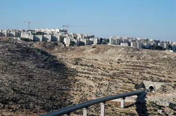 L'image montre un paysage caractérisé par une colline aride et rocheuse, sur laquelle se trouvent des bâtiments modernes. En arrière-plan, on peut voir des constructions en cours, avec des grues qui ajoutent à l'atmosphère de développement urbain. Au premier plan, une structure en béton est visible, semblant faire partie d'une voie ou d'un pont qui traverse le terrain. Le ciel est dégagé, ce qui donne une impression de luminosité et d'espace.