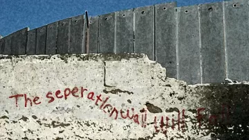 L'image montre un mur ou une barrière en métal, probablement une structure de séparation. À la base, il y a une inscription en rouge qui semble dire "The separation will fall", traduisible en français par "La séparation tombera". Le mur a une texture rugueuse et le contraste entre le métal et le béton met en évidence le message. L'environnement semble désolé, ce qui accentue la portée du message inscrit.