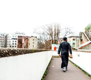 L'image montre un homme marchant sur un chemin urbain. Il porte une tenue traditionnelle, une sorte de longue robe noire. En arrière-plan, on peut voir des bâtiments modernes et anciens, ainsi que des arbres dépouillés. L'environnement semble calme, avec une lumière diffuse, peut-être un jour nuageux. L'homme avance vers l'horizon, de dos, ce qui donne une impression de solitude et de contemplation.