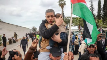 L'immagine mostra un gruppo di persone coinvolte in una manifestazione. In primo piano, un uomo tiene in braccio un bambino, mostrando un'espressione di preoccupazione. Dietro di lui, si vedono altre persone che alzano la voce e agitano bandiere, mentre il contesto sembra essere affollato e vivace. Alcuni partecipanti sono vestiti in modo tradizionale e ci sono alberi e un'architettura più moderna sullo sfondo. L'atmosfera è carica di emozione e impegno.