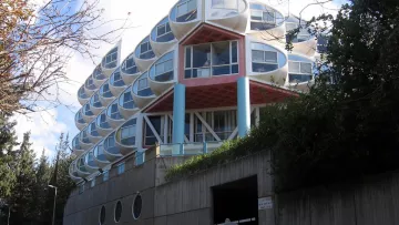 L'image montre un bâtiment moderne avec une architecture unique. Il se compose de plusieurs niveaux, avec une façade caractérisée par des fenêtres en forme de bulle ou de dôme. Les couleurs sont vives, notamment avec des éléments en rouge et en bleu. À l'avant, on peut voir un accès avec un panneau de signalisation et des éléments paysagers comme des arbres sur les côtés. Le bâtiment semble être intégré dans son environnement naturel, avec un design qui attire l'attention.