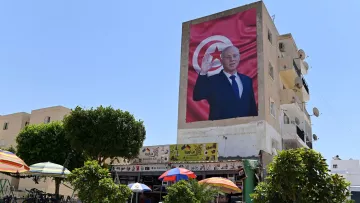 The image features a large mural on the side of a building, depicting a figure in formal attire, raising a hand in a gesture of greeting or salute. The background of the mural includes a red backdrop with a prominent depiction of the national flag of Tunisia. Below the mural, there is a scene of a small open area with umbrellas, likely providing shade for people sitting or gathering. Surrounding the area are some trees and bicycles, suggesting a lively community space. The sky is clear and blue, indicating a bright day.