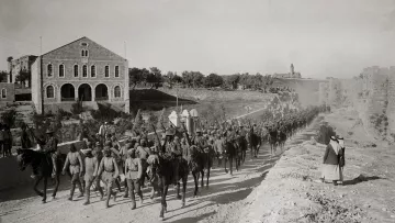 L'image montre une troupe de soldats à cheval marchant en formation le long d'un chemin. En arrière-plan, on peut voir des bâtiments en pierre, dont une grande structure avec des fenêtres. Le sol est poussiéreux, et on remarque également quelques personnes à pied, dont une femme en robe. L'atmosphère semble être celle d'une époque historique, probablement d'un conflit ou d'une quelconque mobilisation militaire. Les soldats portent des uniformes similaires et semblent déterminés dans leur avancée.