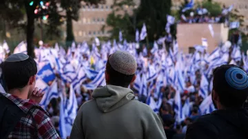 The image shows three individuals, viewed from behind, observing a large crowd at an outdoor event. The crowd is holding blue and white flags, which may symbolize a national identity. The individuals are wearing kippahs (traditional Jewish head coverings). The setting appears to be a public gathering, possibly a rally or demonstration, with trees and buildings visible in the background. The atmosphere seems lively and engaged.