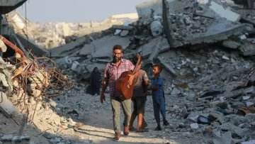 L'image montre un homme marchant à travers des décombres, tenant une guitare. Il est accompagné d'un jeune garçon. L'environnement est en ruines, avec des débris et des structures effondrées autour d'eux. L'atmosphère semble à la fois désolée et résiliente, illustrant un contraste entre la musique et la destruction.