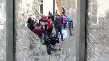 The image shows a group of people gathered on a street, viewed through broken glass. The scene appears to be one of protest or demonstration, as indicated by the presence of individuals wearing masks and the visible graffiti on the walls nearby. Some members of the group are sitting on the ground, suggesting a moment of gathering or reflection amid the chaos. The atmosphere seems tense, reflecting a social or political issue that has drawn this assembly.