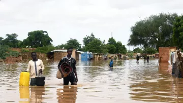 در این تصویر، دو مرد در حال حرکت در یک منطقه‌ی سیلاب‌زده هستند. آب تا زانوهای آنها بالا آمده و چند ساختمان و کلبه‌ در پس‌زمینه به وضوح قابل مشاهده است که بخشی از آنها در آب غرق شده‌اند. آسمان ابری به نظر می‌رسد و تعداد زیادی درخت در اطراف دیده می‌شود. یکی از مردان سطل و دیگری تعدادی وسایل را در دست دارد و به نظر می‌رسد که آنها در تلاش برای عبور از این آب‌ها هستند.