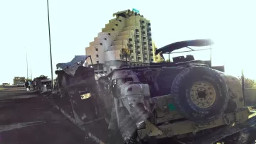 الصورة تظهر مجموعة من المركبات العسكرية المدمرة أو المهجورة على جانب الطريق. في الخلفية، يوجد مبنى شاهق يبدو مهجوراً، ويتميز بتصميمه الفريد. تضيء الشمس الصورة من جهة واحدة، مما يبرز تفاصيل المركبات والمبنى. الجو العام يوحي بالتخلي أو الفوضى، ويعكس مشاهد قد تكون ناتجة عن حالة نزاع أو حرب.
