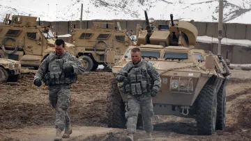 L'image montre deux soldats marchant dans un environnement rocailleux, probablement dans une zone militaire. Ils portent des uniformes camouflage et des gilets tactiques. En arrière-plan, on peut voir des véhicules blindés semblant robustes, probablement utilisés pour des opérations militaires, avec des montagnes enneigées en toile de fond. L'atmosphère semble sérieuse et empreinte d'une certaine rigueur militaire.