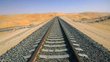 L'image montre une voie de chemin de fer s'étendant à travers un paysage désertique. Les rails, posés sur des traverses en bois, sont entourés de gravier. En arrière-plan, on aperçoit des dunes de sable sous un ciel clair et ensoleillé. Le terrain est principalement aride, et les couleurs dominantes sont des nuances de beige et de gris, ce qui souligne l'isolement et l'immensité de cet environnement désertique.