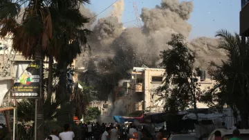 L'image montre une scène chaotique en milieu urbain, où des explosions entraînent un nuage de poussière et de débris. Des bâtiments semblent être endommagés, créant un environnement de destruction. On peut voir des gens dans la rue, réagissant à la situation, ainsi que des palmiers qui ajoutent une touche de verdure au paysage. Le ciel est clair, en contraste avec la fumée épaisse et sombre qui s'élève des ruines.