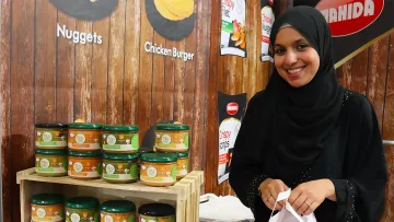 L'image montre une femme portant un hijab, souriant tout en se tenant près d'un stand de produits alimentaires. À côté d'elle, il y a une étagère en bois remplie de pots de produits, probablement des sauces ou des condiments, étiquetés avec différentes saveurs. L'arrière-plan est en bois, et on peut voir d'autres affiches promotionnelles. L'ambiance semble conviviale, et la femme semble enthousiaste à propos des produits exposés.