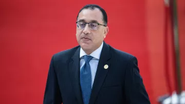 الصورة تظهر رجلًا يرتدي بدلة سوداء مع قميص أبيض ورابطة عنق زرقاء. يبدو أن الرجل في حالة تفكير أو تركيز، حيث تعبير وجهه جدي. الخلفية حمراء، مما يضيف طابعًا رسميًا ومميزًا للصورة.