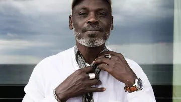 L'immagine ritrae un uomo che indossa una camicia bianca e un cappellino. Ha una barba grigia e un'espressione serena. È circondato da un ambiente luminoso, con una vista sul mare sullo sfondo. L'uomo tiene una sciarpa in mano, mentre i suoi anelli e l'orologio sul polso aggiungono un tocco di stile al suo aspetto. Il cielo appare leggermente nuvoloso, creando un contrasto interessante con l'immagine complessiva.