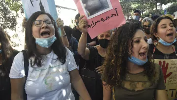 En la imagen se observa a un grupo de personas participando en una protesta. Varias mujeres están al frente, gritando con energía y sosteniendo carteles que muestran fotografías de otras personas. Algunas de las asistentes llevan mascarillas. El ambiente parece lleno de emoción y determinación, con un fondo de árboles y una calle. Las pancartas tienen inscripciones en diferentes idiomas, lo que sugiere que se trata de una manifestación con un propósito importante.