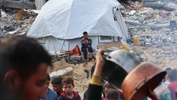 L'image montre un camp de réfugiés après un désastre. Au premier plan, un groupe de personnes autour d'une casserole. En arrière-plan, une tente blanche est visible, avec un jeune assis, regardant son téléphone, tandis que des débris de bâtiments sont éparpillés autour.