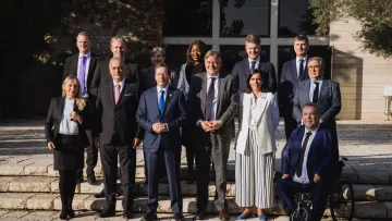 L'image montre un groupe de personnes posant ensemble à l'extérieur, sur des marches devant un bâtiment. Il y a dix-sept individus, dont certains portent des costumes et d'autres des tenues variées. L'arrière-plan est un jardin ou un espace extérieur avec des arbres et une architecture moderne. Les personnes affichent des expressions souriantes, ce qui suggère un moment de camaraderie ou une occasion formelle, comme une réunion ou un sommet.