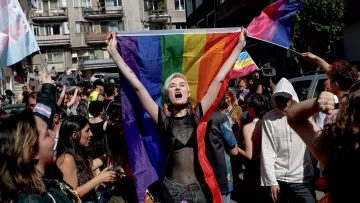 L'immagine mostra un gruppo di persone in una strada affollata, probabilmente durante una manifestazione o una parata. Al centro, una giovane donna con capelli corti e bianchi è in primo piano, mentre tiene alta una grande bandiera arcobaleno. Sembra esprimere gioia e determinazione. Intorno a lei, altre persone sventolano bandiere e manifestano il loro sostegno, creando un'atmosfera di festeggiamento e orgoglio. Le decorazioni e i colori vivaci trasmettono un messaggio di inclusione e solidarietà.
