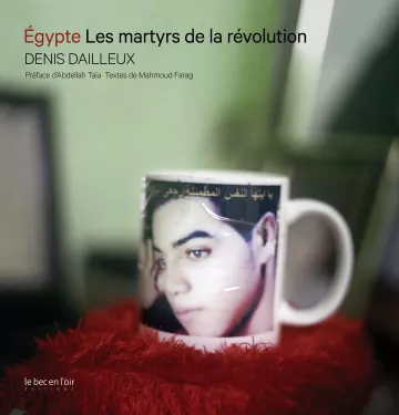 L'image présente une tasse avec une image d'une personne portant un regard triste ou pensif. Le fond de la tasse est légèrement flou, mais on distingue bien le visage. La tasse est placée sur un support de couleur rouge, ajoutant une touche de chaleur à la composition. En bas de l'image, le texte indique "Égypte Les martyrs de la révolution" ainsi que le nom de l'auteur, Denis Dailleux, et des précisions sur la préface et les textes. L'ensemble évoque un hommage ou une réflexion sur les événements révolutionnaires en Égypte.