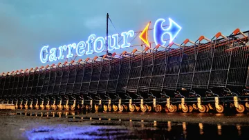 L'image montre un rayonnement lumineux du logo de Carrefour, un hypermarché bien connu, en arrière-plan. Devant le logo, il y a une série de caddies alignés sur le sol, probablement après la pluie, ce qui crée des reflets sur la surface. L'atmosphère est calme et nocturne, accentuée par l'éclairage des enseignes. Les caddies sont organisés de manière ordonnée, offrant une vue intéressante sur l'environnement d'un magasin de grande distribution.