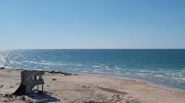 L'image montre une plage sous un ciel bleu clair, avec une mer d'un bleu éclatant qui s'étend à perte de vue. Le rivage est bordé de sable fin, mais des débris, comme une structure en béton en ruine, sont visibles sur la plage, ajoutant un contraste à la beauté naturelle du paysage. Au loin, on peut apercevoir quelques petites embarcations sur l'eau, suggérant une ambiance tranquille et isolée.