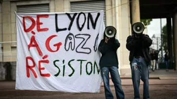 L'immagine mostra due persone che reggono un grande striscione bianco con le parole "DE LYON À GAZA RÉSISTON" scritte in caratteri grandi e colorati. Le due persone hanno in mano dei megafoni e sembrano partecipare a una manifestazione o a un evento di protesta. Lo sfondo suggerisce un contesto urbano, probabilmente in una zona pubblica.