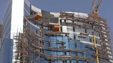 L'image montre un bâtiment en construction. On peut voir une structure moderne avec des façades en verre, entourée d'un échafaudage en métal. Plusieurs niveaux sont visibles, et des éléments de construction, comme des grues, sont présents pour faciliter les travaux. Le ciel est dégagé, ce qui indique une belle journée.