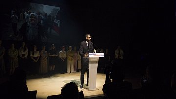 L'immagine mostra un uomo che parla al microfono su un palco, mentre sullo sfondo si vede un'immagine proiettata di una donna con un velo e altre persone. L'oratore indossa un abito elegante e sembra essere il centro dell'attenzione. Dietro di lui, altre persone sono schierate, probabilmente in segno di supporto o partecipazione all'evento. L'illuminazione è drammatica, creando un'atmosfera intensa e concentrata sull'oratore.