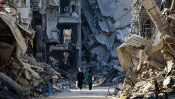 L'image montre un paysage de destruction urbaine, avec des bâtiments effondrés sur les côtés d'une rue. On peut apercevoir deux personnes marchant au milieu des décombres. L'atmosphère est sombre et désolée, avec des ruines qui témoignent d'une catastrophe récente. Les déchets et les restes de matériaux de construction jonchent le sol, créant un sentiment de désolation.