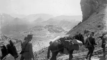 L'image montre un paysage aride et montagneux, avec des soldats marchant à pied, accompagnés de chevaux chargés de matériel. Les soldats portent des uniformes et se déplacent sur un chemin sinueux en direction de formations rocheuses impressionnantes. L'atmosphère est celle d'une expédition militaire dans une région difficile d'accès. Les montagnes en arrière-plan ajoutent une dimension majestueuse et austère à la scène.